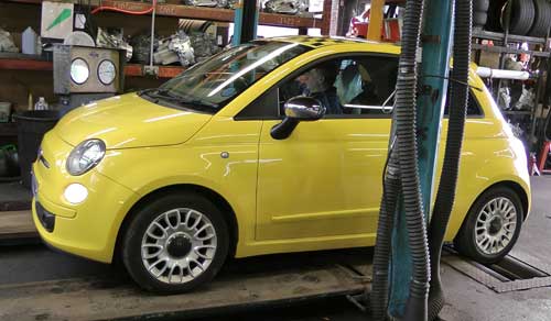 front passenger side distance view of a really pretty bright yellow fiat 500 getting its annual mot test on the rolling brake test with our mot inspector at the wheel